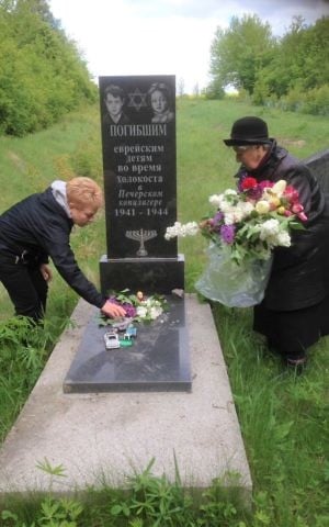 Holocaust survivor Rita Schweibes places flowers on a memorial to child Holocaust victims in Tulchyn, Ukraine, May 10, 2017. Mass burial graves can be seen behind. (Sue Surkes)