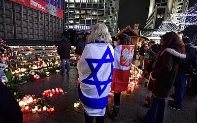 Women covered with Israeli (L) and Polish flags (R)  pay their respect at the memorial shift for the victims of the 2016 deadly truck attack at the Christmas market at Breitscheidplatz in front of the Kaiser-Wilhelm-Gedaechtniskirche during the commemorations marking one year after it occurred, on December 19, 2017 in Berlin. (AFP PHOTO / Tobias SCHWARZ)