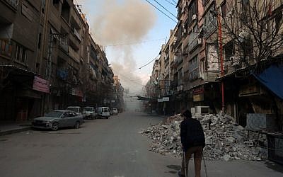 A Syrian man on crutches walks down a street as smoke billows in the rebel-held town of Douma, in the besieged Eastern Ghouta region on the outskirts of the capital Damascus, following airstrikes by regime forces on the area on February 23, 2018. (AFP/ HAMZA AL-AJWEH)