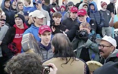 In this January 18, 2019 image made from video provided by the Survival Media Agency, a teenager wearing a "Make America Great Again" hat, center left, stands in front of an elderly Native American singing and playing a drum in Washington. (Survival Media Agency via AP)