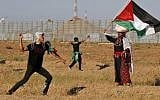 A Palestinian rioter hurls stones at Israeli troops as a woman wearing a traditional Palestinian outfit waves a national flag near the border with Israel, east of Gaza City, on May 10, 2019. (Photo by Said KHATIB / AFP)