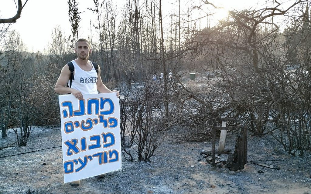A resident stands next to burned grounds in Mevo Modi'im holds a sing that reads "Mevo Modiim Refugee Camp" in protest of the government's lack of support for the moshav, May 30, 2019. (courtesy Inbal Mustacchi) A resident stands next to burned grounds in Mevo Modi'im holds a sing that reads "Mevo Modiim Refugee Camp" in protest of the government's lack of support for the moshav, May 30, 2019. (courtesy Inbal Mustacchi)