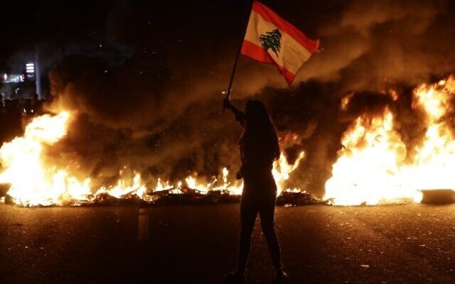 A Lebanese anti-government protester waves a Lebanese flag in front of burning tires that block the main highway linking the city of Tripoli to Beirut at the coastal city of Byblos, north of the capital on November 13, 2019. (Photo by Joseph EID / AFP) A Lebanese anti-government protester waves a Lebanese flag in front of burning tires that block the main highway linking the city of Tripoli to Beirut at the coastal city of Byblos, north of the capital on November 13, 2019. (Photo by Joseph EID / AFP)