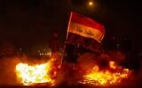 An Iraqi anti-government protester holds a national flag as he stands by a fire at a makeshift roadblock in the southern city of Basra late on December 25, 2019. (Photo by Hussein FALEH / AFP)