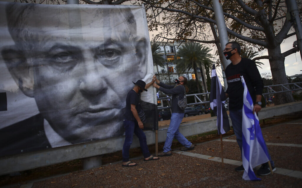 In this Saturday, April 25, 2020 photo, demonstrators wearing protective face masks amid concerns over the country's coronavirus outbreak, hang a banner showing Israeli Prime Minister Benjamin Netanyahu during "Black Flag" protest against Netanyahu and government corruption, at Rabin Square in Tel Aviv, Israel.  (AP Photo/Oded Balilty)