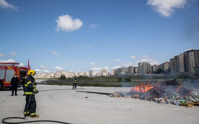 Israeli firefighters at a ceremony burning chametz, or leaven products, collected by the Jerusalem municipality in a final preparation before the Passover holiday, near the disused runway at the abandoned Atarot Airport, north of Jerusalem, on April 8, 2020. In the background are high-rises in the Kafr Aqab Palestinian neighborhood -- an area formally part of municipal Jerusalem but beyond the security barrier. (Yonatan Sindel/Flash90) Israeli firefighters at a ceremony burning chametz, or leaven products, collected by the Jerusalem municipality in a final preparation before the Passover holiday, near the disused runway at the abandoned Atarot Airport, north of Jerusalem, on April 8, 2020. In the background are high-rises in the Kafr Aqab Palestinian neighborhood -- an area formally part of municipal Jerusalem but beyond the security barrier. (Yonatan Sindel/Flash90)