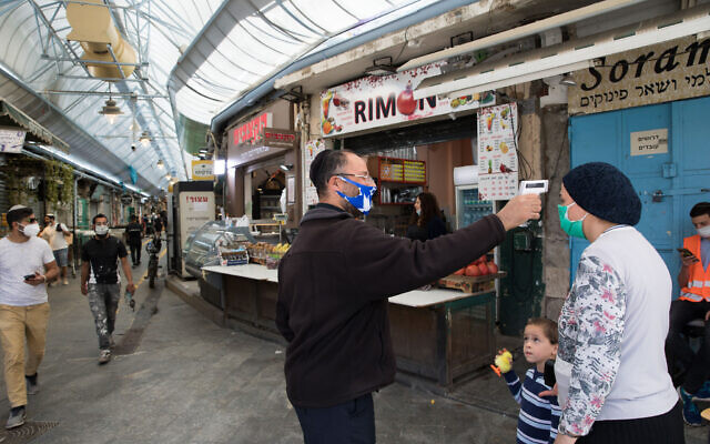 A man takes the temperature of a customer to check if she has a fever, at an entrance to the Mahane Yehuda market in Jerusalem, May 04, 2020. (Nati Shohat/Flash90) A man takes the temperature of a customer to check if she has a fever, at an entrance to the Mahane Yehuda market in Jerusalem, May 04, 2020. (Nati Shohat/Flash90)