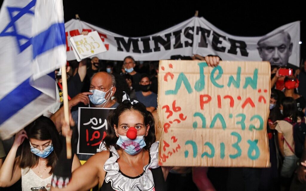 An anti-government protester dressed as a clown and wearing a mask due to the coronavirus pandemic raises a sign reading 'The emergency government is a sad joke' during a demonstration in Charles Clore Park in Tel Aviv on July 18, 2020. (Jack Guez/AFP)
