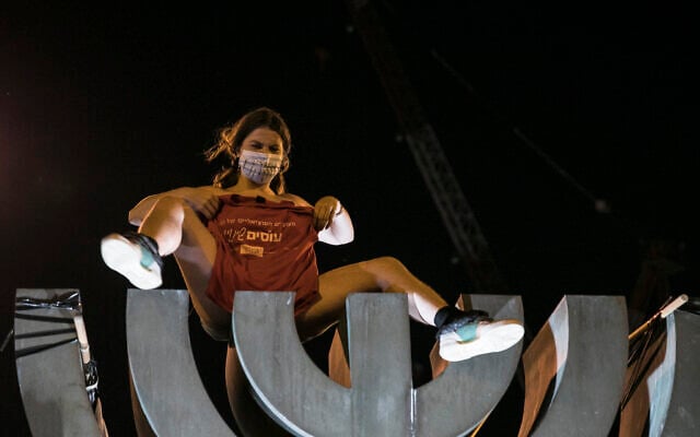 A topless protester sits on a menorah statue near the Knesset during a demonstration against Prime Minister Benjamin Netanyahu in Jerusalem on July 21, 2020. (Yonatan Sindel/Flash90) A topless protester sits on a menorah statue near the Knesset during a demonstration against Prime Minister Benjamin Netanyahu in Jerusalem on July 21, 2020. (Yonatan Sindel/Flash90)