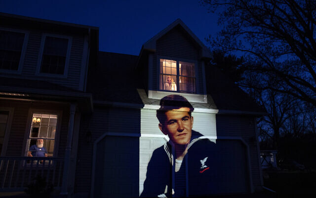 An image of veteran Stephen Kulig is projected onto the home of his daughter, Elizabeth DeForest, as she looks out the window of a spare bedroom as her husband, Kevin, sits downstairs in Chicopee, Mass., Sunday, May 3, 2020. Kulig, a US Navy veteran and resident of the Soldier's Home in Holyoke, Mass., died from the COVID-19 virus at the age of 92. After saying goodbye to her father for the last time in person, Elizabeth slept in the spare bedroom upstairs for two weeks as a precaution against possibly infecting her husband. Seeking to capture moments of private mourning at a time of global isolation, the photographer used a projector to cast large images of veterans on to the homes as their loved ones are struggling to honor them during a lockdown that has sidelined many funeral traditions. (AP/David Goldman)