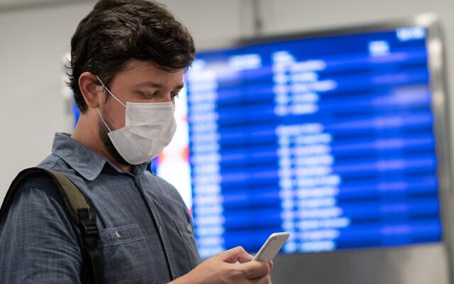 A mask-wearing traveler looks at his cellphone in front of a flight panel (Vergani_Fotografia; iStock by Getty Images) A mask-wearing traveler looks at his cellphone in front of a flight panel (Vergani_Fotografia; iStock by Getty Images)