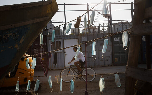 A man wearing face mask rides his bicycle next to boats with face masks hanging on them during a nationwide lockdown to curb the spread of the coronavirus, at Jaffa port near Tel Aviv, October 7, 2020. (Oded Balilty/AP) A man wearing face mask rides his bicycle next to boats with face masks hanging on them during a nationwide lockdown to curb the spread of the coronavirus, at Jaffa port near Tel Aviv, October 7, 2020. (Oded Balilty/AP)