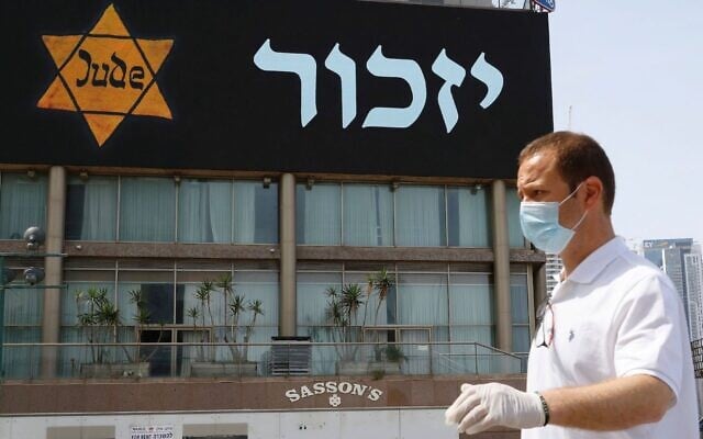 Illustrative: A man in a mask and protective gear stands near a banner depicting a Holocaust-era yellow Star of David, in Tel Aviv on April 21, 2020. (Jack Guez/AFP via Getty Images) Illustrative: A man in a mask and protective gear stands near a banner depicting a Holocaust-era yellow Star of David, in Tel Aviv on April 21, 2020. (Jack Guez/AFP via Getty Images)