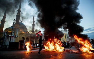 Mounir Hujairi, 23, holds up a Lebanese national flag as walks in front of burning tires set on fire to block a main road, during a protest in downtown Beirut, Lebanon, March 3, 2021. (AP Photo/Hassan Ammar, File)