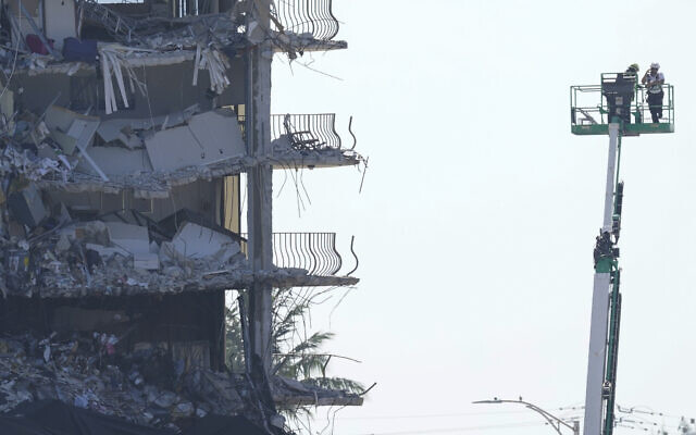 Workers stand watch from a lift beside the partially collapsed Champlain Towers South condo building, where demolition experts were preparing to bring down the precarious still-standing portion, Sunday, on July 4, 2021, in Surfside, Florida. (AP/Lynne Sladky) Workers stand watch from a lift beside the partially collapsed Champlain Towers South condo building, where demolition experts were preparing to bring down the precarious still-standing portion, Sunday, on July 4, 2021, in Surfside, Florida. (AP/Lynne Sladky)