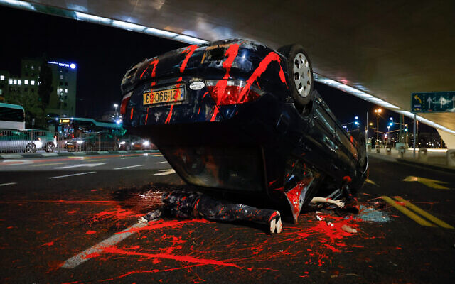 A flipped-over car painted with fake blood, with a demonstrator lying beneath the overturned vehicle, in an exhibit accompanying a nationalist protest demanding a probe of the death of teenager Ahuvia Sandak last year, at the entrance to Jerusalem, November 21, 2021. (Olivier Fitoussi/Flash90) A flipped-over car painted with fake blood, with a demonstrator lying beneath the overturned vehicle, in an exhibit accompanying a nationalist protest demanding a probe of the death of teenager Ahuvia Sandak last year, at the entrance to Jerusalem, November 21, 2021. (Olivier Fitoussi/Flash90)
