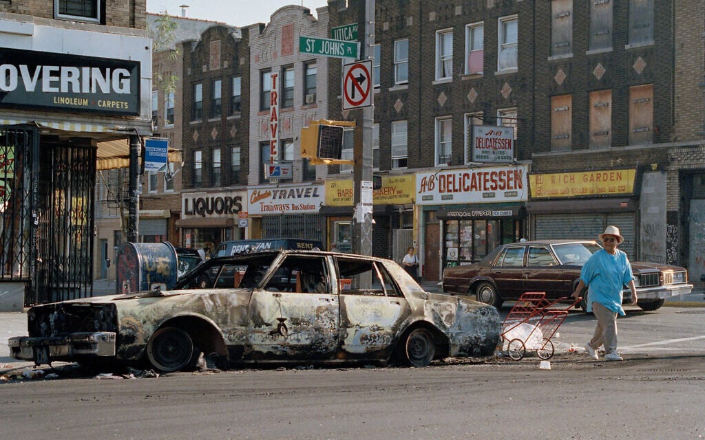 FILE - In this August 22, 1991 file photo, a woman passes a fire-bombed car as she crosses St. Johns Place on Utica Avenue in the Crown Heights section of the Brooklyn borough of New York. (AP Photo/David Cantor, File)