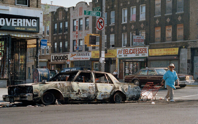 FILE - In this August 22, 1991 file photo, a woman passes a fire-bombed car as she crosses St. Johns Place on Utica Avenue in the Crown Heights section of the Brooklyn borough of New York. (AP Photo/David Cantor, File)