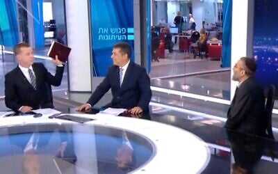 Political commentator Amit Segal, left, raises a copy of Maimonides' 'Hilchot Talmud Torah' during an interview with United Torah Judaism leader Moshe Gafni, right, as his co-host Ben Caspit smiles, on Channel 12's 'Meet the Press' on September 17, 2022. (Screen capture)