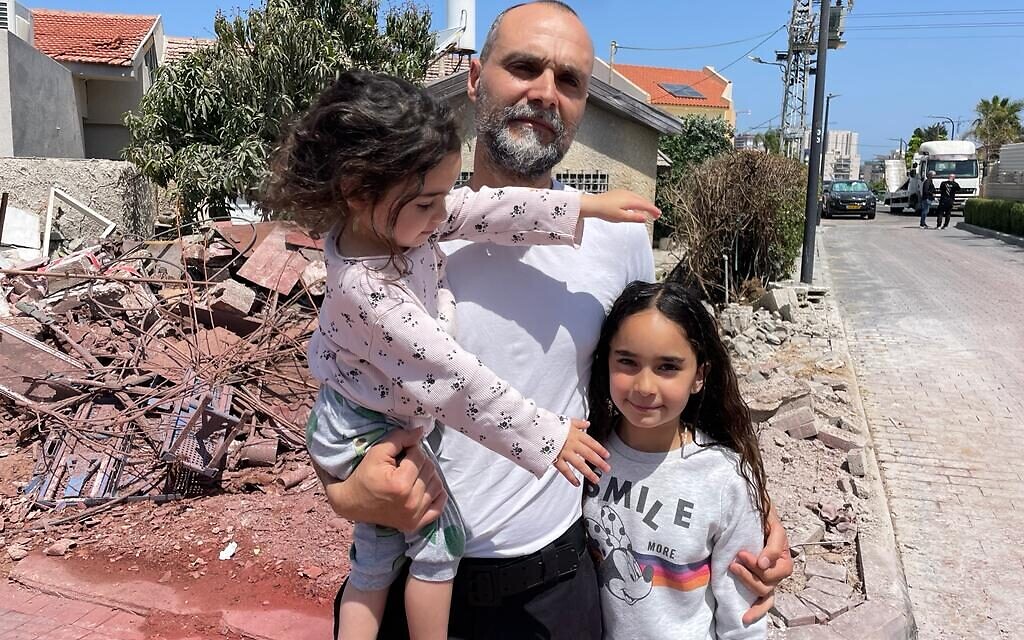 Nissim Azoulai and his two daughters stand in front of the wreckage of a direct rocket hit in their Ashkelon neighborhood, May 11, 2023 (Carrie Keller-Lynn/The Times of Israel)