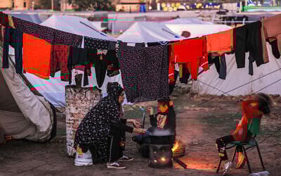 A woman sits by food cooking on a fire as children play nearby at the southern Gaza Strip on December 18, 2023. (Mahmud Hams / AFP)