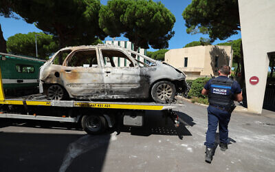 A police officer walks past a burnt car being moved on the bed of a towing vehicle in the seaside resort of La Grande Motte, near the city of Montpellier, southern France, on August 26, 2024, following the August 24, 2024 attack to the Beth Yaacov synagogue. (Pascal GUYOT / AFP)