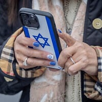 Illustrative: A woman holds her cellphone with an Israeli flag cover at the Mahane Yehuda Market in Jerusalem, February 23, 2024. (Chaim Goldberg/Flash90)