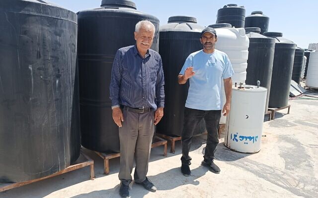 Two residents of the Jerusalem neighborhood of Kafr Aqab stand next to tanks placed by local residents on rooftops and filled with water purchased privately, as their water supply is cut to only a few hours a week, July 24, 2024 (Gianluca Pacchiani/Times of Israel)