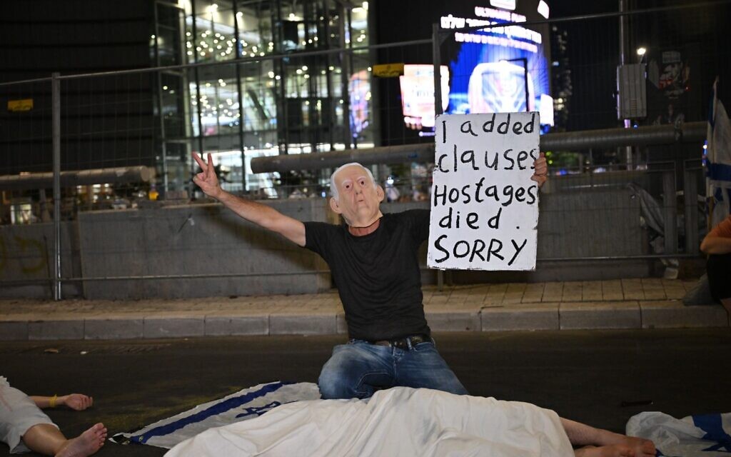 A protester wearing a mask of Prime Minister Benjamin Netanyahu holds a sign that says, "I added clauses [to the hostage deal] and hostages died. Sorry," while kneeling over mock bodies of dead hostages, at Tel Aviv's Begin Gate on August 31, 2024. (Neta Dekel/Pro-Democracy Protest Movement)