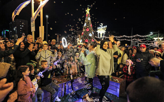 Girls pose for pictures on a platform as bubbles are sprayed at them near a lit up Christmas tree, Ramadan Lantern, and Hanukkah menorah displayed at the German Colony area in Israel's northern city of Haifa on December 21, 2024. (Ahmad Gharabli / AFP)