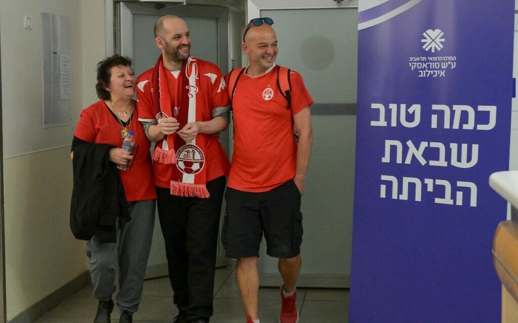 Newly-released hostage Iair Horn (center), wearing a Hapoel Beersheba soccer shirt, flanked by his mother and brother, pictured at Tel Aviv Sourasky Medical Center on February 15, 2025. (Ma'ayon Toaf / GPO)