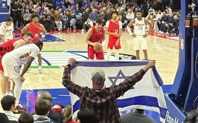Fan Lavi Kirel waves an Israeli flag as basketball player Deni Avdija takes a free throw during a March 3 game in Philadelphia. (Jon Marks/Times of Israel)