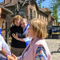 Holocaust survivor Irene Shashar (R) speaks to rescued hostage Ori Megidish (L), while Holocaust survivor Gita Kaufman and released hostage Agam Berger embrace, draped in an Israeli flag outside the "Arbeit Macht Frei" sign at Auschwitz during March of the Living on April 24, 2025 in Oswiecim, Poland. (Alexi Rosenfeld/Getty Images, via JTA)
