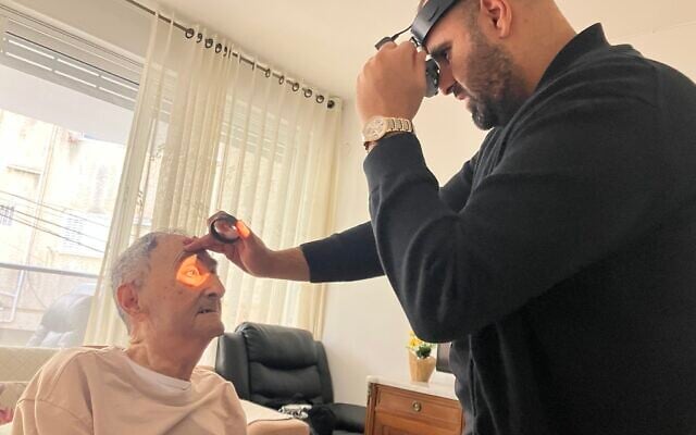 Dr. Fadi Badarna, right, a volunteer eye doctor with  LeMa’anam (“for their sake” in Hebrew), a non-profit organization providing timely healthcare free of charge to Holocaust survivors in their homes, examines Nissim Daito's eyes at his house in Kiryat Bialik on April 16, 2025. (Diana Bletter/Times of Israel)