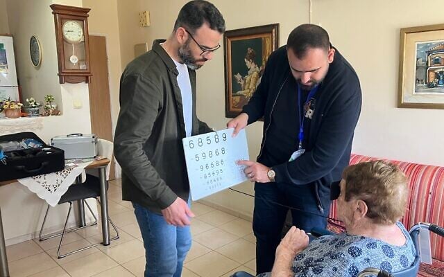Dr. Yosif Boulos, left, and Dr. Fadi Badarna, volunteer eye doctors with  LeMa’anam (“for their sake” in Hebrew), a non-profit organization providing timely healthcare free of charge to Holocaust survivors in their homes, give an eye examination to Chana Sandouvsky in her Kiryat Bialik apartment on April 16, 2025. (Diana Bletter/Times of Israel)