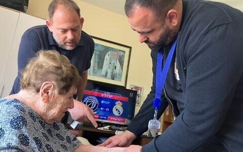 Dr. Fadi Badarna, right, a volunteer eye doctor with LeMa’anam (“for their sake” in Hebrew), a nonprofit organization providing free home healthcare to Holocaust survivors, and Aviv Paz, a LeMa'anam employee, look at the number tattoo that Chana Sandouvsky received in Auschwitz, in her home in Kiryat Bialik on April 16, 2025. (Diana Bletter/Times of Israel)