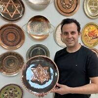 Dror Miro, a fourth-generation coppersmith, holds his 'Rising Lion' plate he designed and made in the old city of Acre on June 16, 2025. (Diana Bletter/Times of Israel)