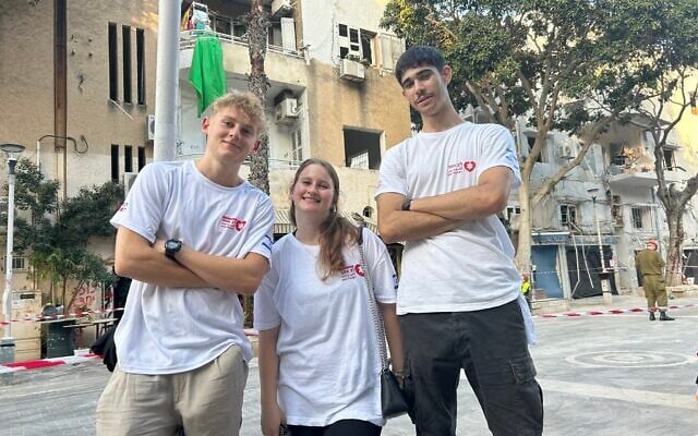 Lev Echad volunteers (from left) Ray Margolin, May Esher, and Eyal Meidan, stand against a background of buildings damaged by an Iranian missile on June 22, 2025. (Sue Surkes/Times of Israel)