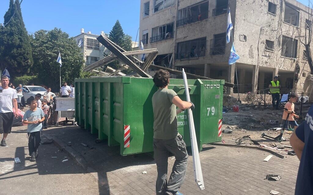 A volunteer throws parts of a blind damaged by an Iranian missile explosion into a dumpster in the northern city of Haifa, June 16, 2025. (Sue Surkes/Times of Israel)