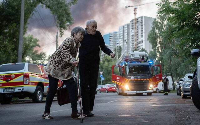An elderly couple crossing a street as smoke rises in the background at a site of a strike following a mass Russian drone and missile attack on the Ukraine's capital Kyiv, July 4, 2025. (Handout / STATE EMERGENSY SERVICE OF UKRAINE / AFP)