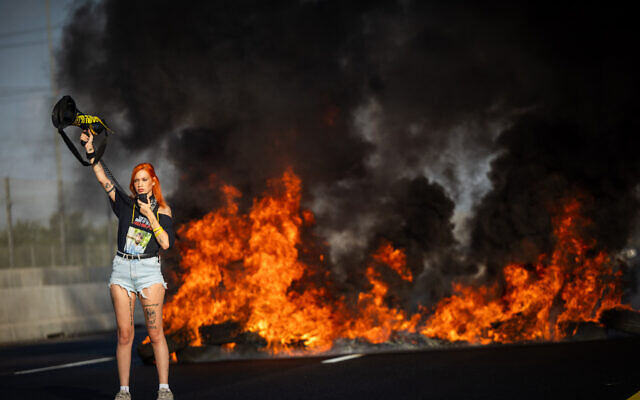 A protester blocks a main road near Lod as a fire blazes behind her, during a demonstration calling for the release of the hostages held by Hamas and a ceasefire in Gaza, August 26, 2025.  (Yonatan Sindel/Flash90)