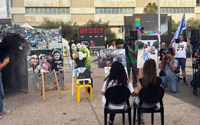 Arbel Yehoud and Sharon Aloni-Cunio sit facing photos of their partners, brothers Ariel and David Cunio, in an installation at Tel Aviv's Hostages Square on August 26, 2025. (Ariela Karmel/Times of Israel)