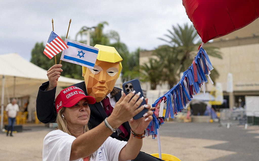 A person wearing a mask depicting US President Donald Trump holds US and Israeli flags at Hostages Square in Tel Aviv on October 9, 2025. (Maya Levin / AFP) A person wearing a mask depicting US President Donald Trump holds US and Israeli flags at Hostages Square in Tel Aviv on October 9, 2025. (Maya Levin / AFP)
