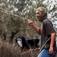 A masked Jewish extremist swings a slingshot while hurling stones at Palestinians who had gathered for the annual olive harvest season, during an attack by Israeli settlers on the Palestinian village of Beita, south of Nablus in the West Bank, on October 10, 2025. (Photo by Jaafar ASHTIYEH / AFP)