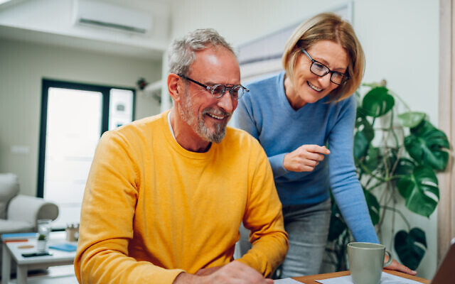Senior middle aged happy couple using laptop together at home. Smiling elderly family reading news or shopping online. Older people and technology concept. Focus on a man in a yellow sweater.

JCF