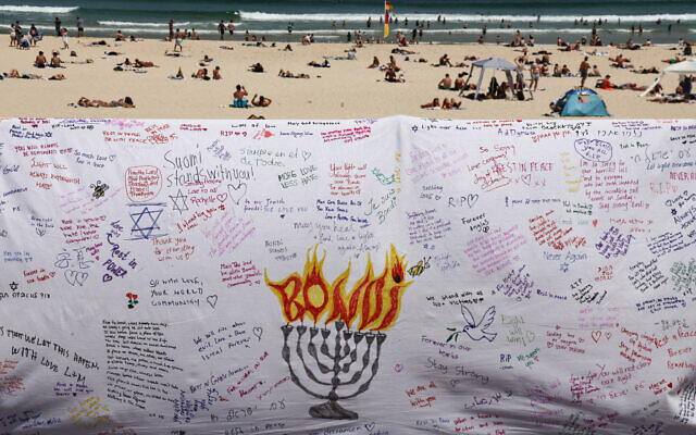 Beachgoers are seen behind a piece of cloth bearing messages left by mourners at the promenade of Bondi Beach in Sydney on December 18, 2025, to honor the victims of the terror attack that targeted a Jewish community event marking the first night of Hanukkah on December 14. (David Gray / AFP) Beachgoers are seen behind a piece of cloth bearing messages left by mourners at the promenade of Bondi Beach in Sydney on December 18, 2025, to honor the victims of the terror attack that targeted a Jewish community event marking the first night of Hanukkah on December 14. (David Gray / AFP)