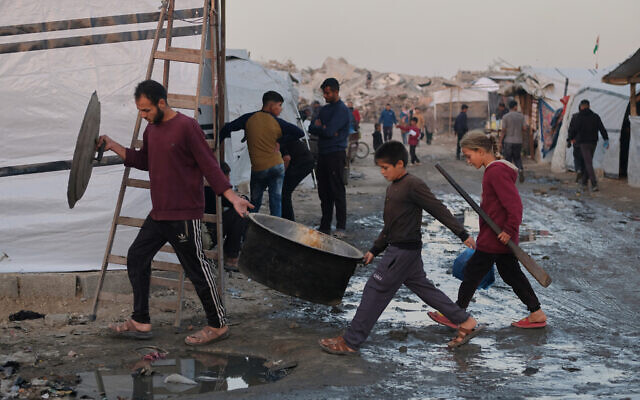 Displaced Palestinians walk through muddy pathways as a man and children carry cooking equipment at a camp in Jabalia, in the northern Gaza Strip, Dec. 7, 2025. (AP Photo/Jehad Alshrafi)