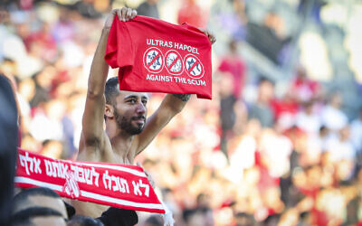 A Hapoel Tel Aviv fan holds up a controversial t-shirt reading "Against the scum," with the logos of the Israel Police, the banned Kach party, and Maccabi Tel Aviv, at the Bloomfield Stadium in Tel Aviv, December 20, 2025. (Danny Maron/Flash90)