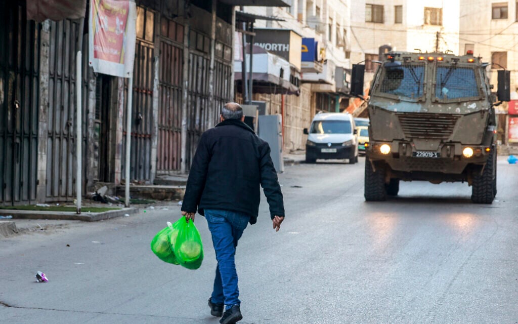 A Palestinian man carries food as an Israeli military vehicle drives past shuttered shops during a Israeli military operation in the old city of the northern West Bank city of Nablus on January 11, 2026. (Jaafar ASHTIYEH / AFP)