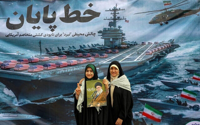 Women pose with a poster depicting Iran's Supreme Leader Ayatollah Ali Khamenei as they stand before a banner depicting a US Navy aircraft carrier with US-flag-wrapped coffins on the deck, during a rally marking the 47th anniversary of the 1979 Islamic revolution in Tehran on February 11, 2026 (AFP)